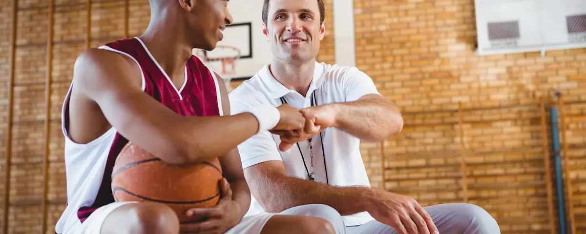 Basketball player in uniform holds ball and fist bumps mentor in a gym, showing supportive athletic program relationships