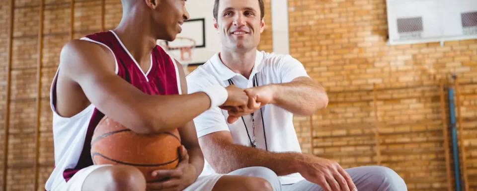 Basketball player in uniform holds ball and fist bumps mentor in a gym, showing supportive athletic program relationships