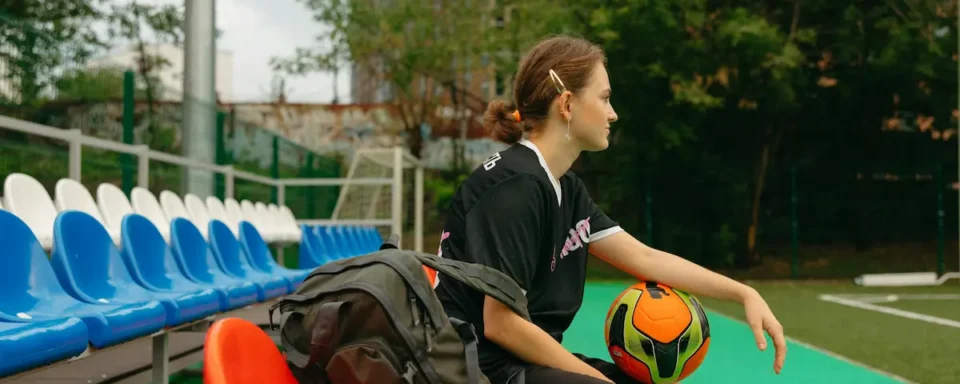 An athlete sitting on a bench, holding their backpack and soccer ball on their lap.