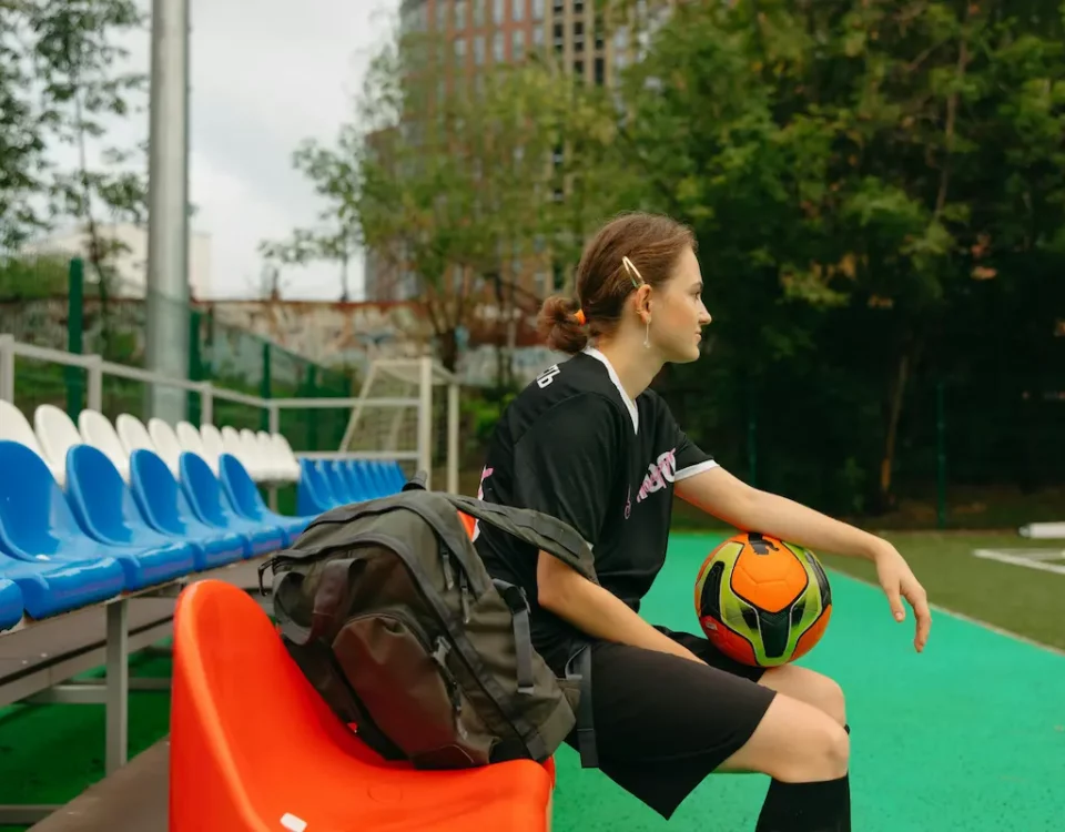 An athlete sitting on a bench, holding their backpack and soccer ball on their lap.