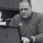 Athletic director in a school works on a laptop at his office desk, preparing for game day.
