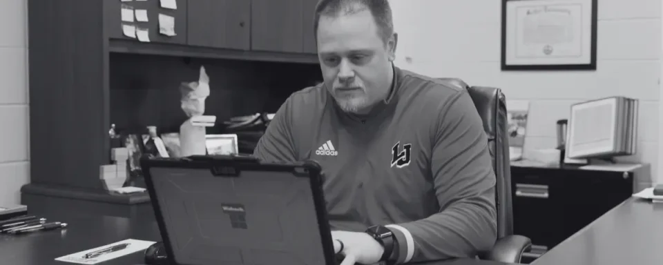 Athletic director in a school works on a laptop at his office desk, preparing for game day.