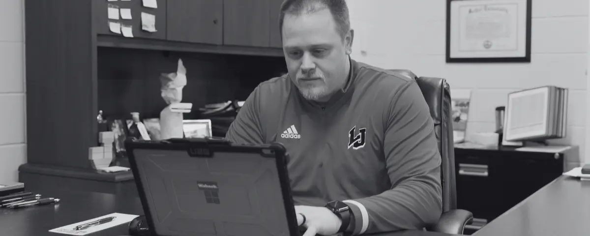 Athletic director in a school works on a laptop at his office desk, preparing for game day.
